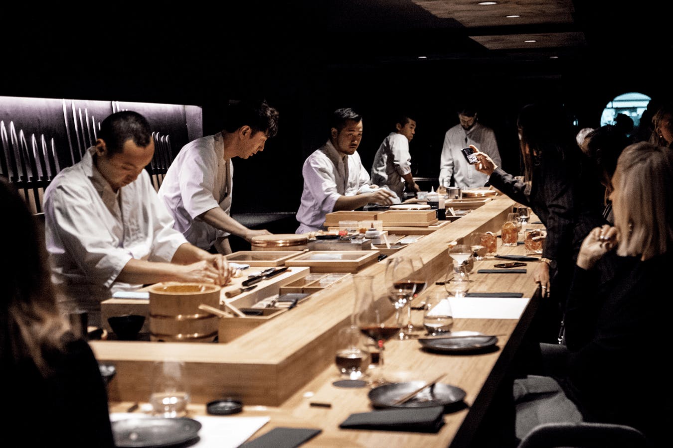 A group of chefs crafting sushi at a wooden bar with diners sitting watching.